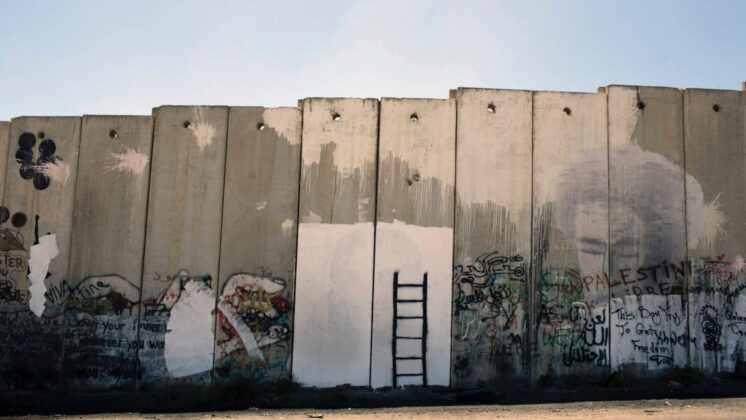 Photo of annexation wall on Palestinian side. Wall is covered with graffiti, notably including a black ladder on a white background. The ladder does not reach the top of the wall